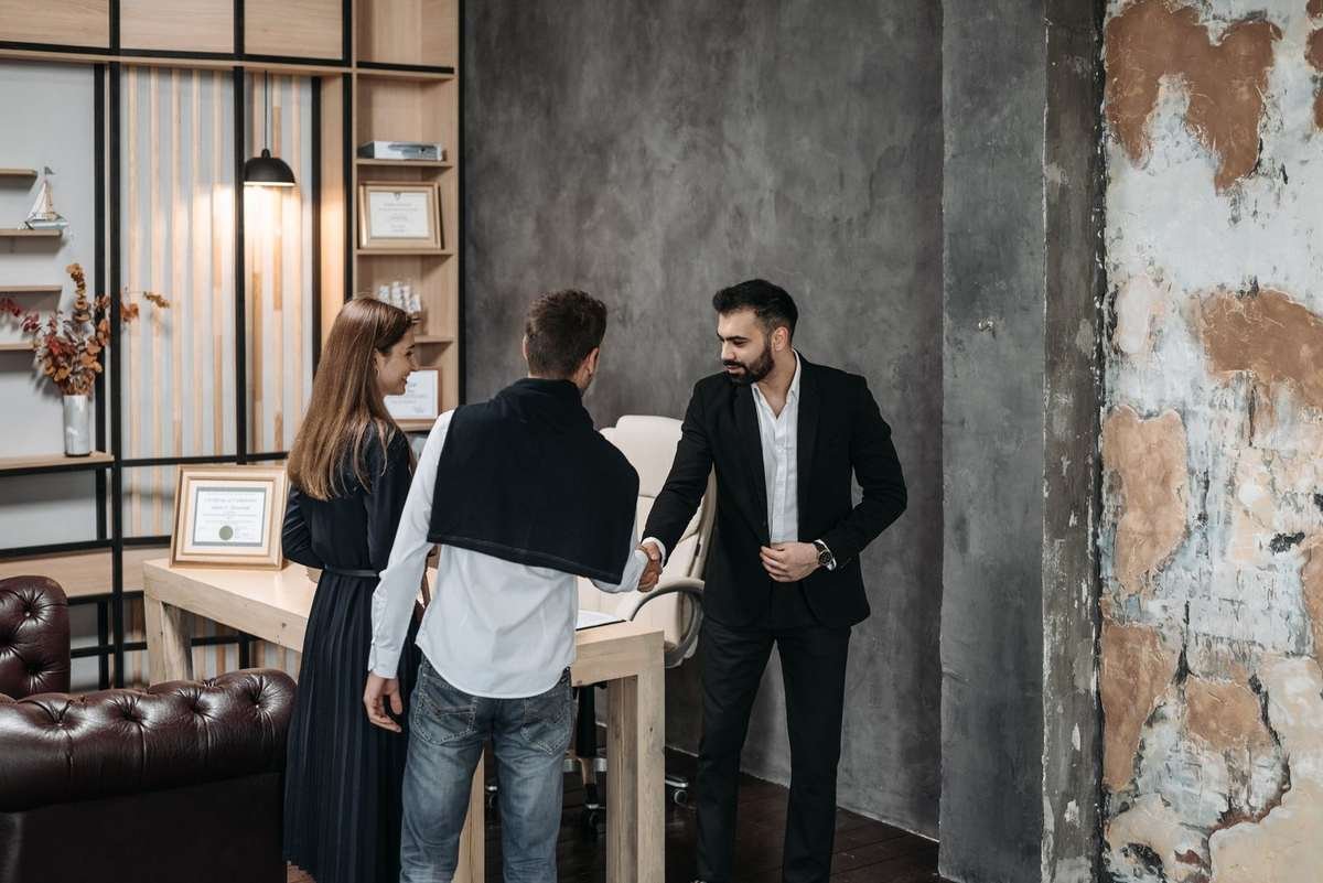 Homme en costume noir serre la main d'un autre homme, femme en noir à côté, salon avec bureau et lampe Homme en costume noir serre la main d'un autre homme, femme en noir à côté, salon avec bureau et lampe