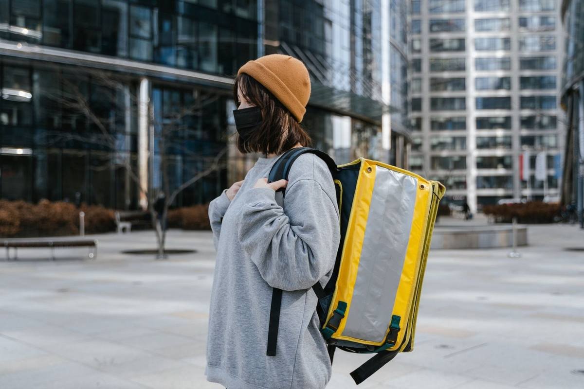 Femme portant un sac jaune et un masque, vêtue d'un manteau gris, dans un environnement urbain avec des bâtiments modernes