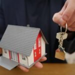Une main tient une petite maison modèle et une clé sur une table