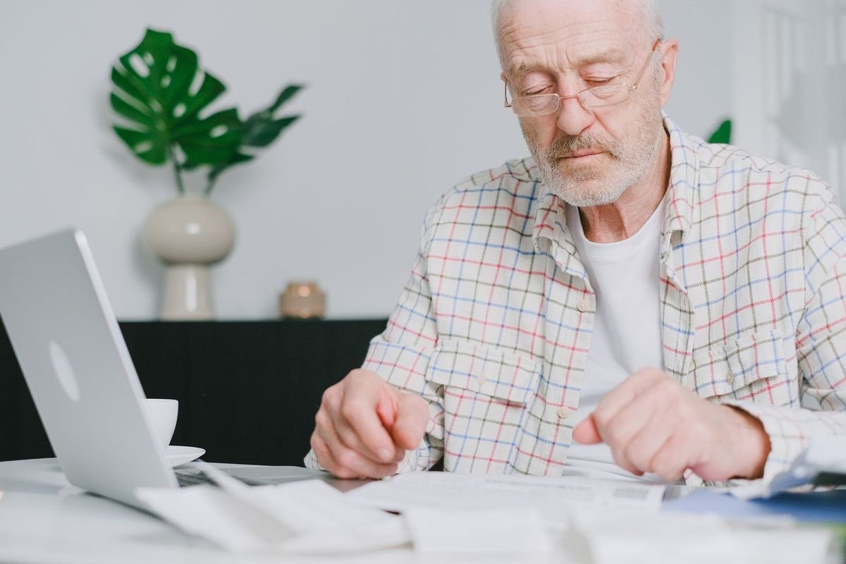 Homme âgé devant un ordinateur portable, en train de travailler sur des papiers, à l'intérieur d'une maison Homme âgé devant un ordinateur portable, en train de travailler sur des papiers, à l'intérieur d'une maison