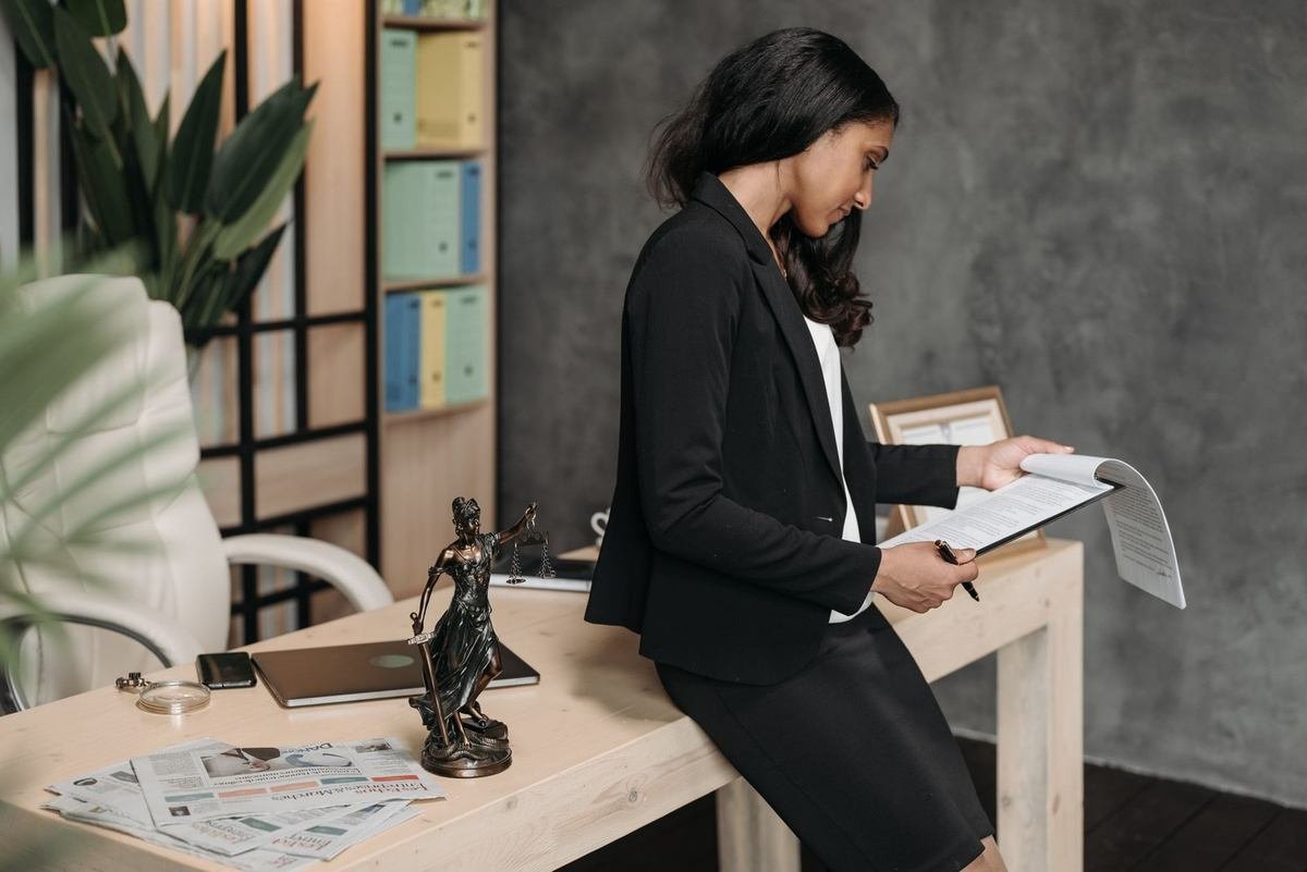Femme en costume noir lit un document à un bureau en bois, avec une statue, un ordinateur et des papiers