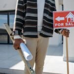 Homme avec planche à niveau et rouleau de papier devant maison, panneau "À vendre" à la main