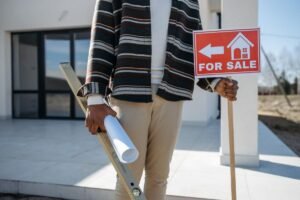 Homme avec planche à niveau et rouleau de papier devant maison, panneau "À vendre" à la main